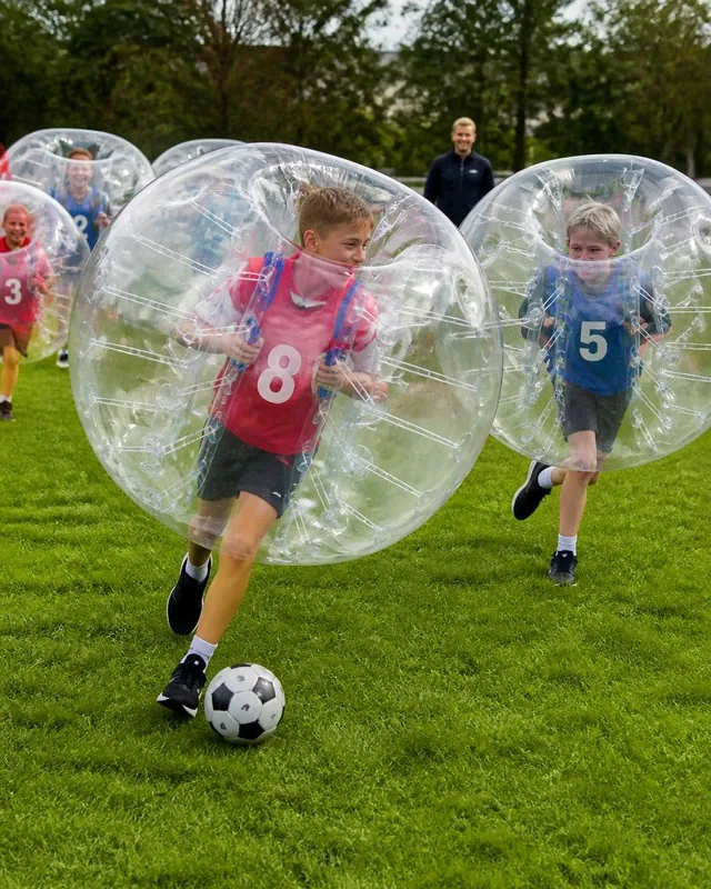 Barn i röda och blå västar jagar en fotboll med bumperballs på gräs.