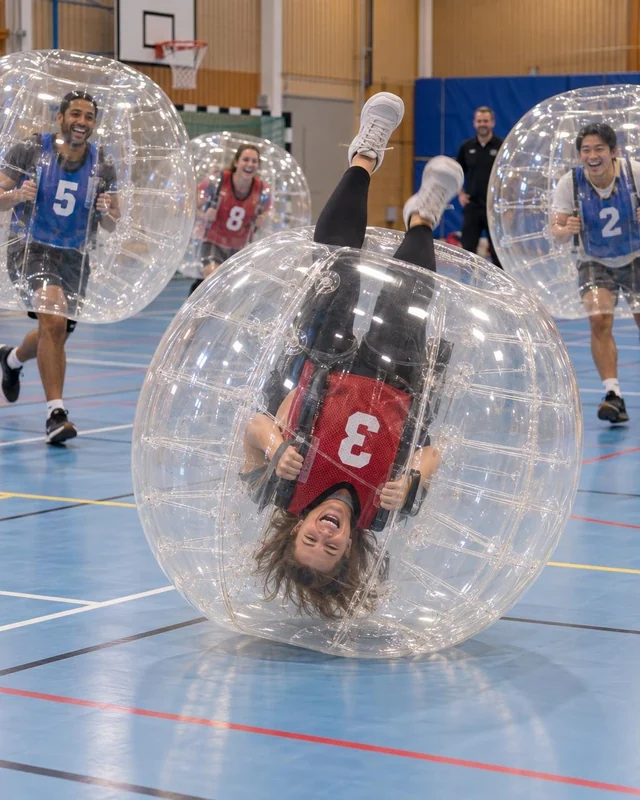 En vuxen i röd väst är upp och ned i en bumperball på blått sporthallsgolv.