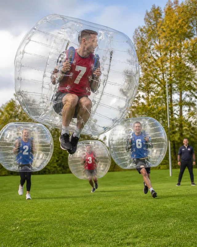 En vuxen i röd väst hoppar i en bumperball medan andra spelare ser på.