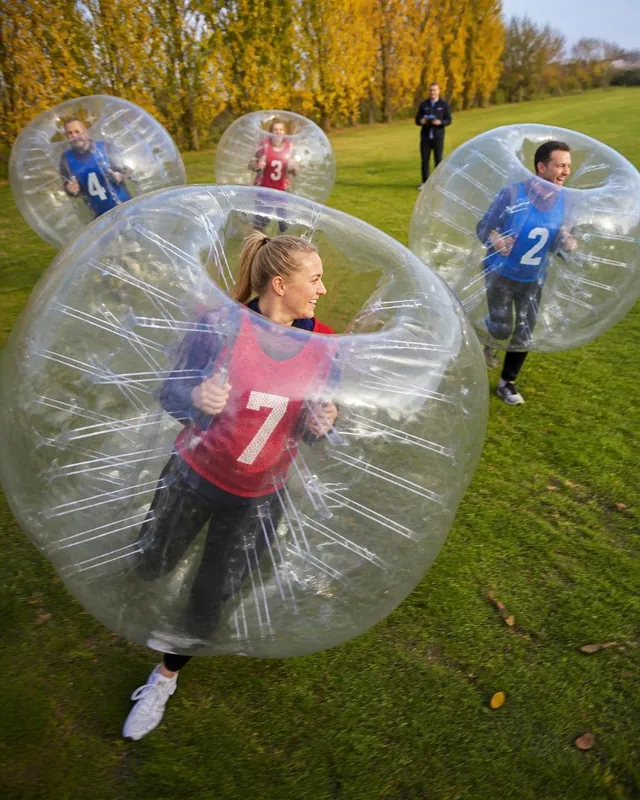 Vuxna spelar med genomskinliga bumperballs på gräs framför gula höstträd.